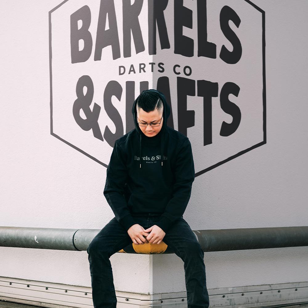 Sweat à capuche Barrels and Shafts - Noir Eine Person trägt einen schwarzen "Barrels and Shafts" Hoodie und sitzt auf einer Bank. Im Hintergrund ist ein großes Wandlogo mit der Aufschrift "Barrels & Shafts Darts Co" zu sehen.