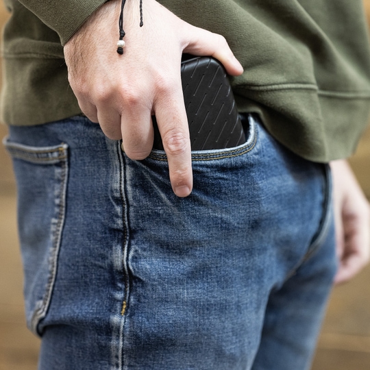 Custodia per freccette Harrows Slimcase Eine Person steckt ein schwarzes Etui in die Hosentasche. Sie trägt eine grüne Jacke und eine blaue Jeans.
