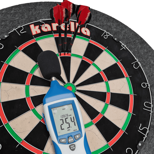 811501_Karella_Schallschutz_f-r_Steeldartboards_40AsdKuqQ3Dv9I Das Bild zeigt ein Steeldartboard mit integriertem Schallschutz und Auffangring von Karella. Auf dem Dartboard liegen drei rote Darts sowie ein Schallpegelmessgerät, das 25,4 dB anzeigt.
