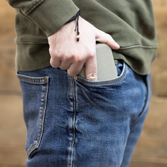 Etui na lotki Harrows Slimcase Eine Person steckt eine graue Darttasche in ihre Hosentasche. Die Person trägt eine blaue Jeans und einen olivgrünen Pullover.