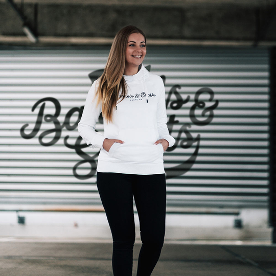 Barrels and Shafts Damen Hoodie - Weiß Eine Frau trägt einen weißen "Barrels and Shafts" Damen Hoodie. Sie steht vor einem Hintergrund mit großem Schriftzug und lächelt.
